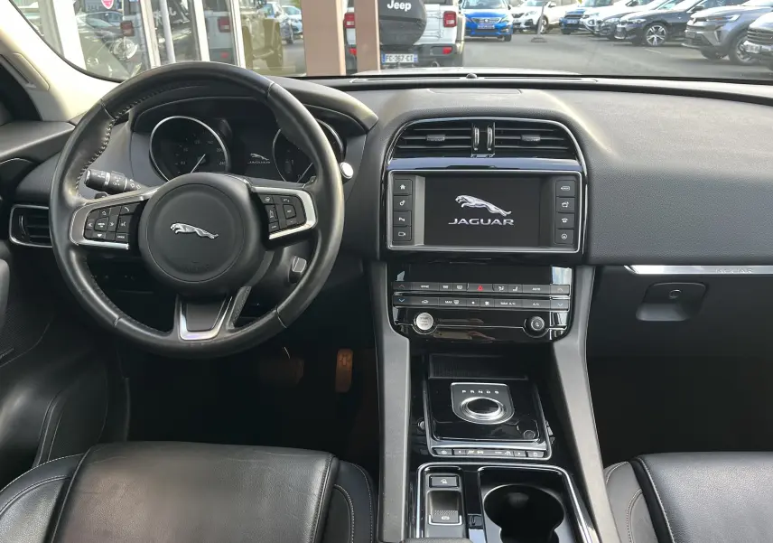 Vue intérieure avant du Jaguar F-PACE blanc 2018, tableau de bord avec écran tactile et volant cuir noir multifonction.
