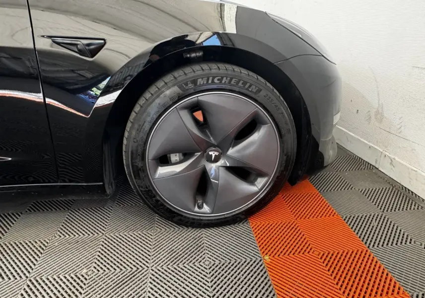 Gros plan sur la roue avant gauche noire d'une Tesla Model 3 2023 avec jante aérodynamique et logo Tesla central.