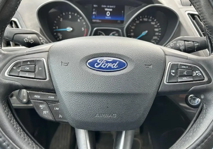 Vue rapprochée du volant cuir noir avec logo Ford et commandes intégrées du tableau de bord d'une Ford C-MAX gris clair.