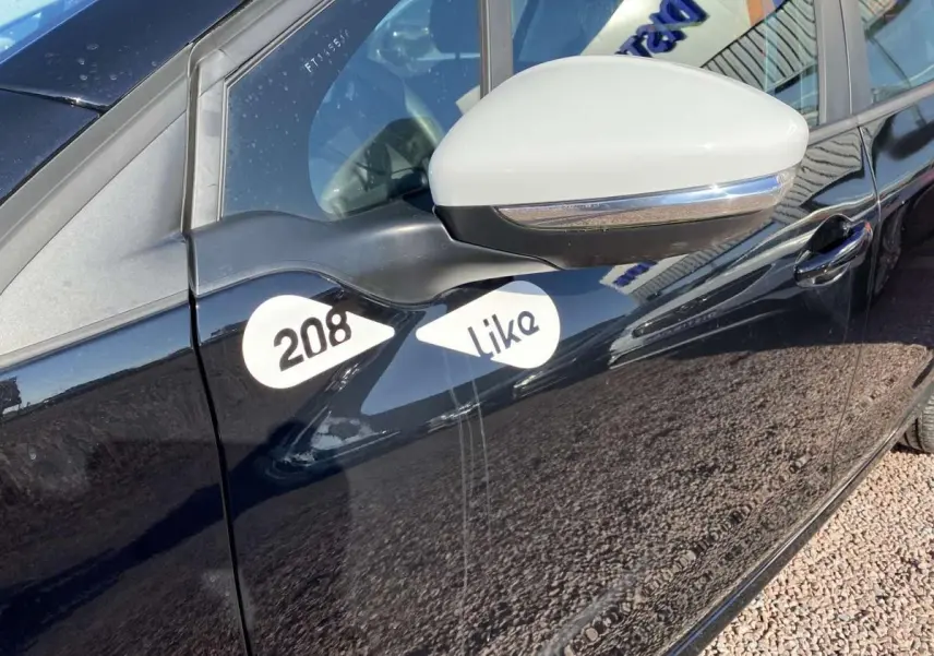 Vue côté gauche rapprochée d'une Peugeot 208 noire avec coques de rétroviseurs blanches et stickers "208 Like" sur la porte.