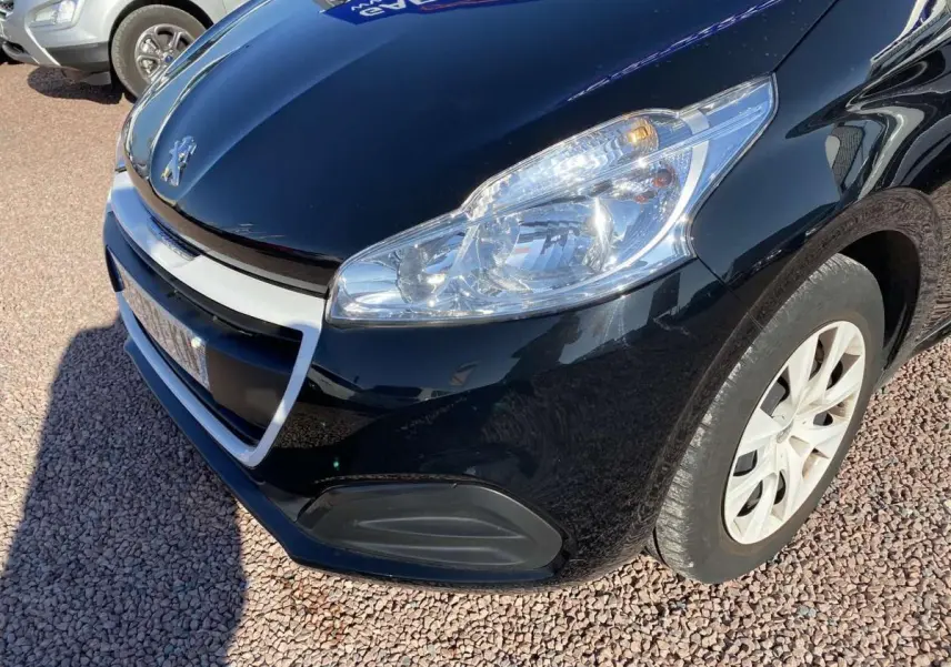Vue en 3/4 avant droit d'une Peugeot 208 noire avec jonc de calandre blanc et phare allumé sur sol gravillonné.