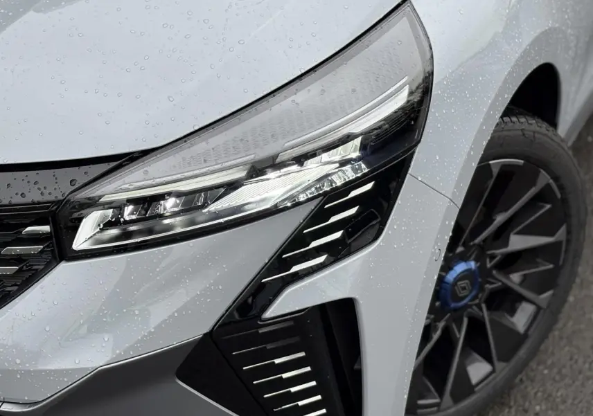 Gros plan sur le phare avant droit et la jante alliage noire avec cabochon bleu d'une Renault Clio Gris Rafale 2025.