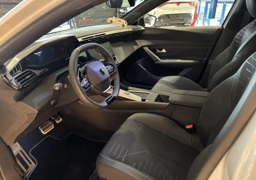 Vue intérieure côté conducteur de la Peugeot 308 blanche, avec volant compact GT et sièges tissu noir à surpiqûres vertes.