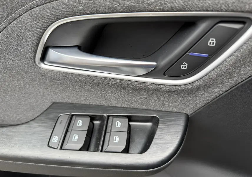 Poignée de porte intérieure et commandes des vitres en tissu gris et plastique noir du MG ZS 1.5L Hybrid+ 2026 côté conducteur.