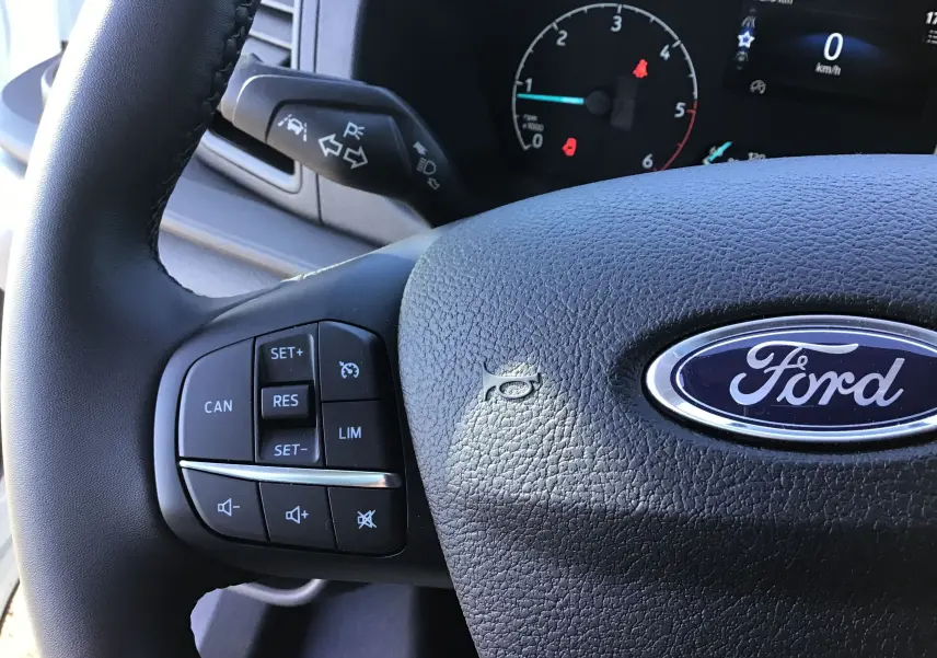 Gros plan sur le volant noir du Ford Transit Châssis Cabine blanc, avec commandes intégrées et logo Ford visible.