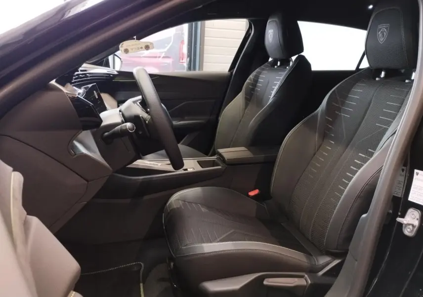 Vue intérieure côté conducteur de la Peugeot 308 noir métal, sièges tissu noir avec logo Peugeot brodé sur appuie-tête.