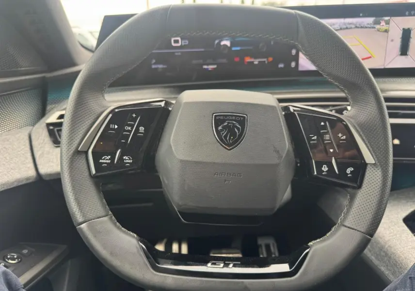 Vue rapprochée du volant noir du Peugeot 5008 Hybrid GT 2025 avec commandes intégrées et logo central.