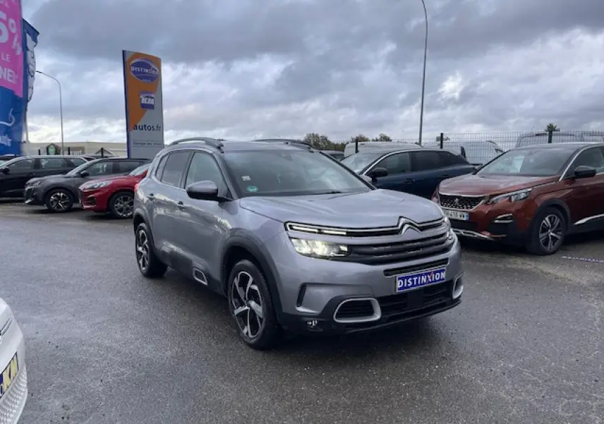 Citroën C5 Aircross gris Artense vue 3/4 avant avec feux allumés sur parking sous ciel nuageux.