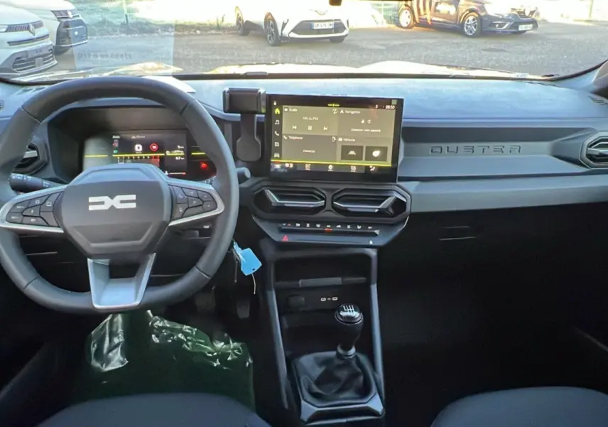 Vue intérieure du tableau de bord du Dacia Duster 2025, avec volant multifonction, écran tactile et levier de vitesse manuel.