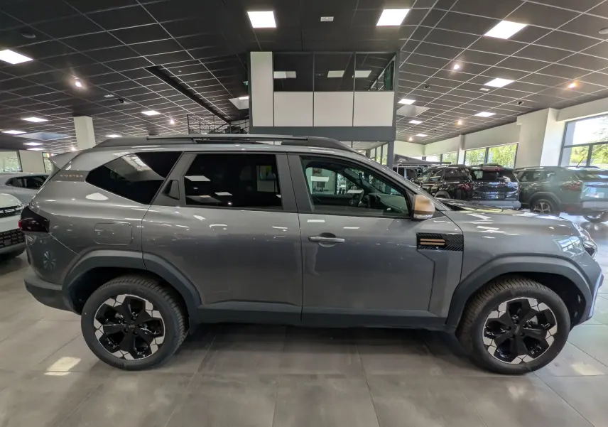 Vue de profil côté droit du Dacia Bigster Hybrid-G 150 4x4 gris schiste avec jantes biton noires en showroom.