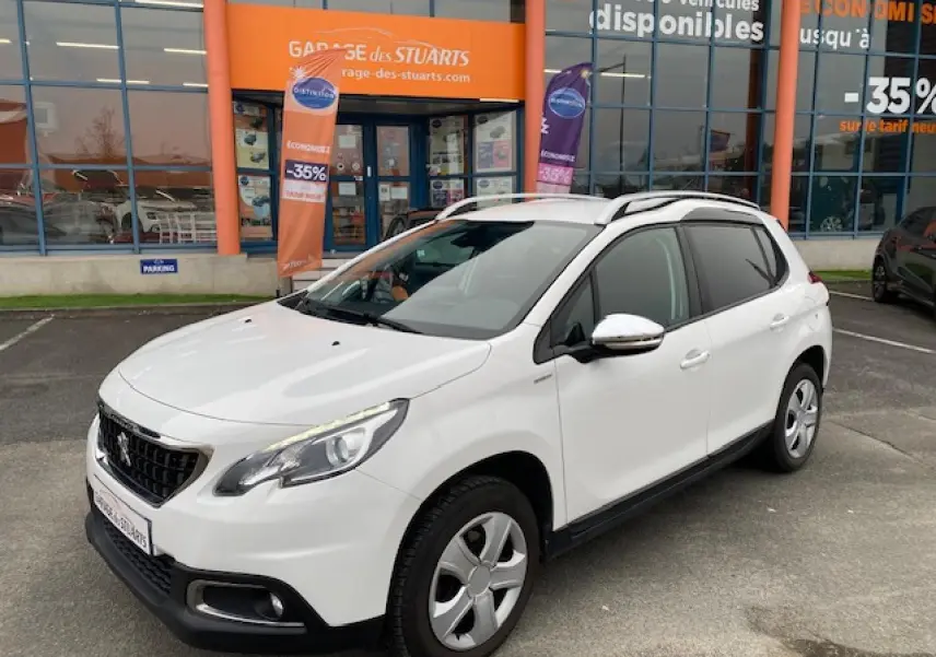 Peugeot 2008 blanc vue 3/4 avant droit, avec jantes acier et barres de toit, stationné devant un garage orange.