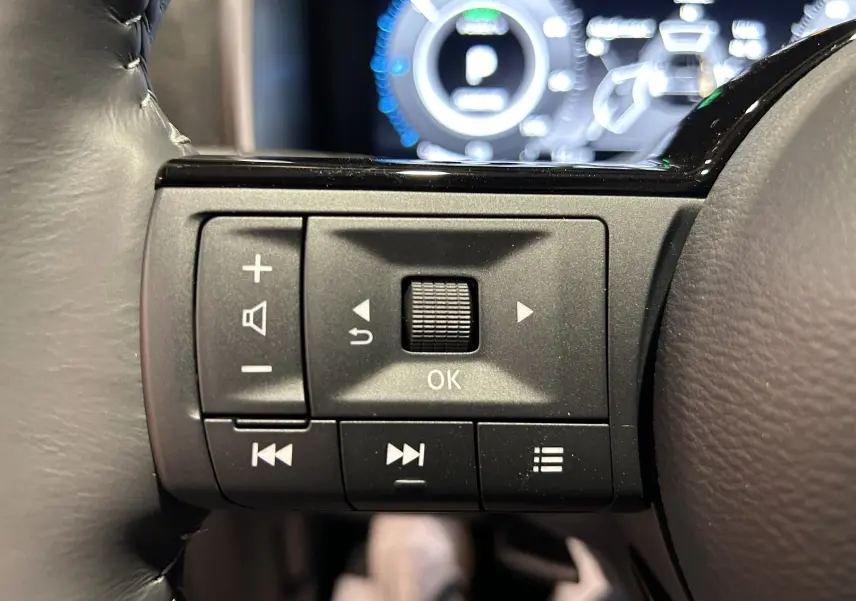 Gros plan sur les commandes multifonctions noires du volant du Nissan Qashqai 2025, avec tableau de bord numérique flou en arrière-plan.
