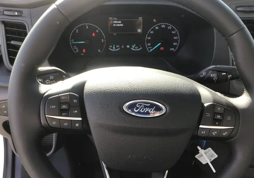Vue rapprochée du volant noir Ford avec commandes intégrées et tableau de bord numérique allumé du Transit Chassis Cabine blanc.