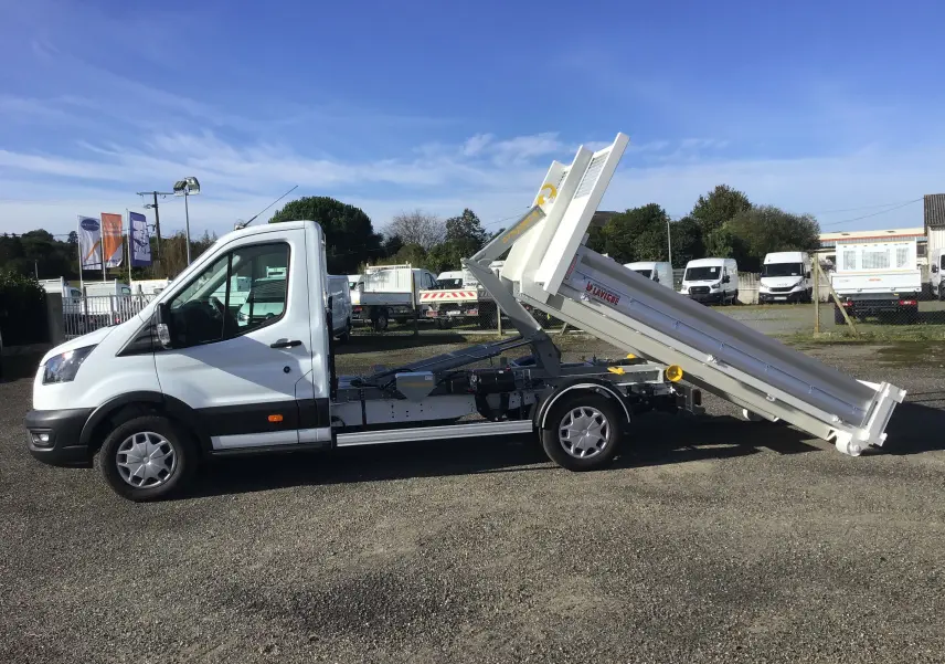 Vue de profil côté gauche d'un Ford Transit blanc avec benne relevée et bras polybenne visible sur un parking extérieur.