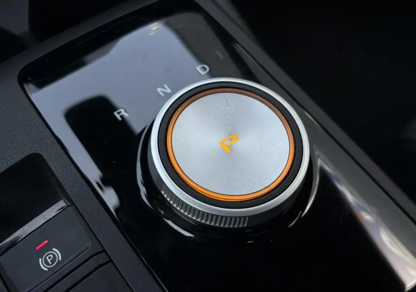 Gros plan sur la molette de sélection de boîte automatique argentée avec éclairage orange dans l’habitacle noir du MG3 blanc.