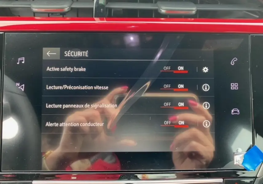 Écran tactile 7 pouces de l'Opel Corsa 2022 affichant les options de sécurité activées, reflet de mains avec ongles rouges.