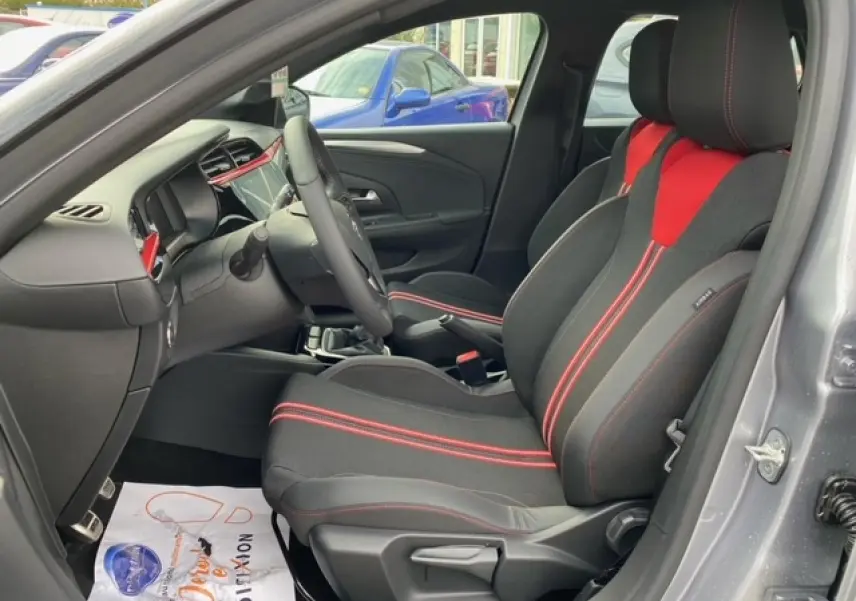 Intérieur de l’Opel Corsa gris magnetic, vue côté gauche, mettant en valeur les sièges noirs avec surpiqûres rouges et tableau de bord moderne.