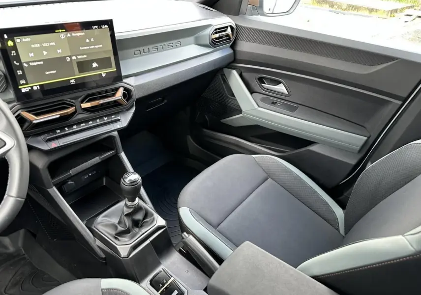 Vue intérieure côté passager du Dacia Duster 2025, tableau de bord noir avec écran tactile et levier de vitesses manuel.