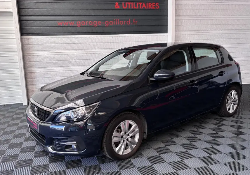 Peugeot 308 gris foncé vue 3/4 avant droit, stationnée en intérieur sur sol damier avec enseigne rouge en arrière-plan.