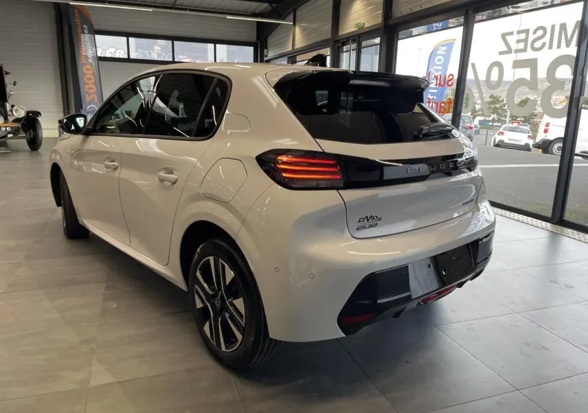 Vue 3/4 arrière droite d'une Peugeot 208 blanche Okenite avec bandeau noir brillant et feux arrière à LED en intérieur showroom.