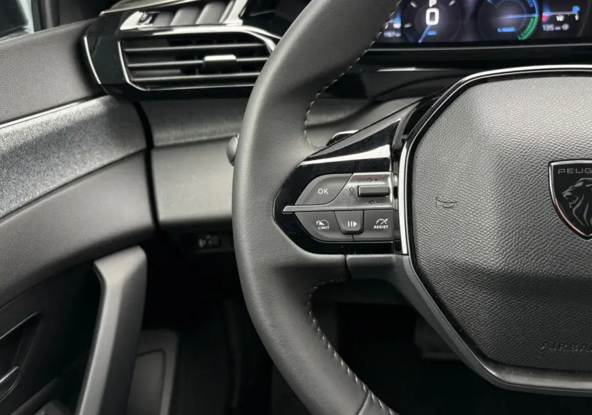 Gros plan sur le volant noir de la Peugeot 308 SW Hybrid 2025 avec commandes intégrées et tableau de bord numérique partiel.