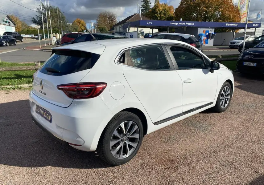 Renault Clio blanche vue 3/4 arrière droit, avec jantes alliage et vitres teintées sur parking extérieur.