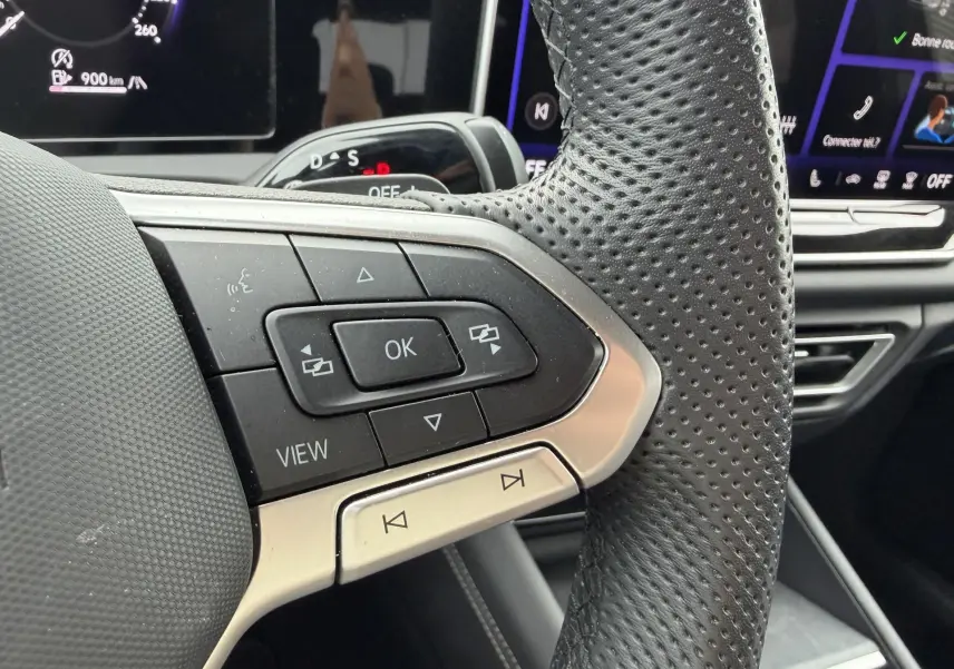 Gros plan sur les commandes gauche du volant en cuir perforé du Volkswagen Tiguan gris, avec écran numérique en arrière-plan.