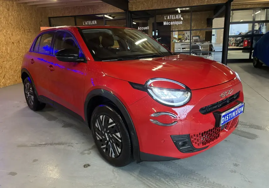 Vue 3/4 avant droite d'une FIAT 600 1.2 Hybrid 2024 rouge avec phares allumés et jantes stylisées en intérieur d'atelier.