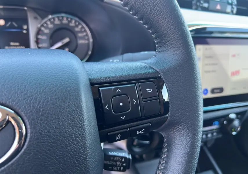 Gros plan sur le volant cuir multifonctions noir du Toyota Hilux 2026 avec écran tactile et tableau de bord flou en arrière-plan.