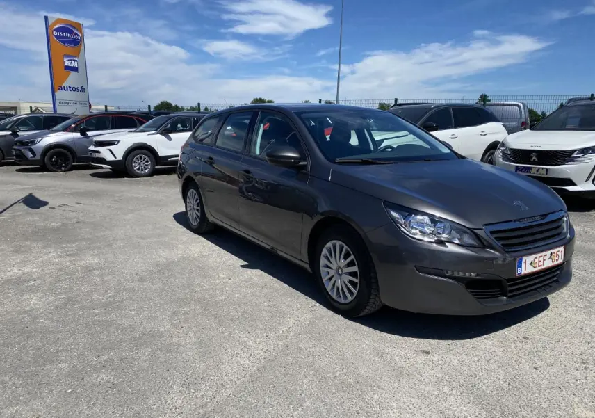 Peugeot 308 SW gris clair vue 3/4 avant droit sur parking ensoleillé avec attelage visible à l'arrière.