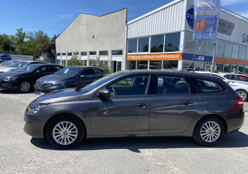Profil côté gauche d'une Peugeot 308 SW gris clair garée devant un bâtiment commercial sous un ciel bleu.