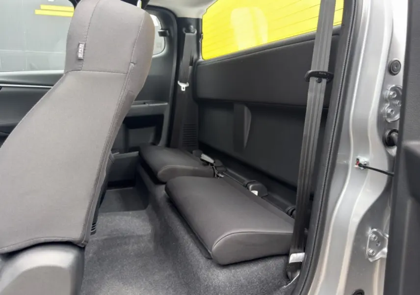 Vue intérieure côté droit du pick-up Isuzu D-MAX 2026, sièges arrière rabattus en tissu noir, carrosserie argentée visible.