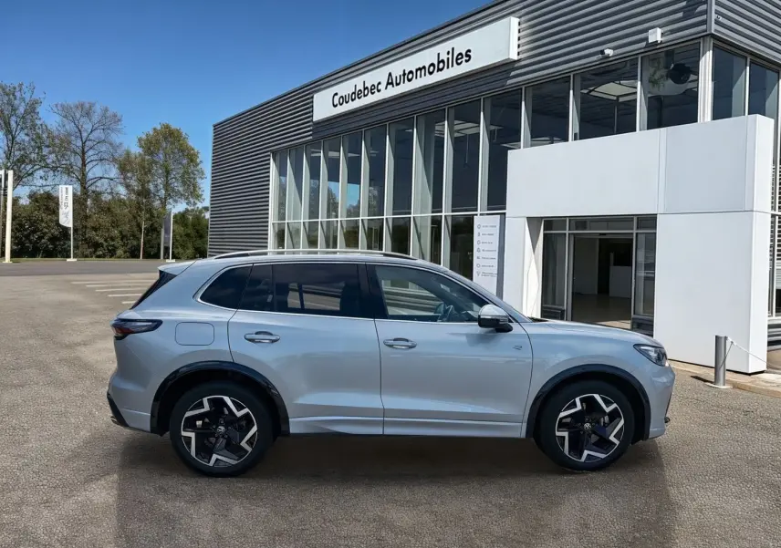 Vue latérale droite du Volkswagen Tiguan gris 2025 avec jantes alliage noires et toit ouvrant, devant un bâtiment moderne.
