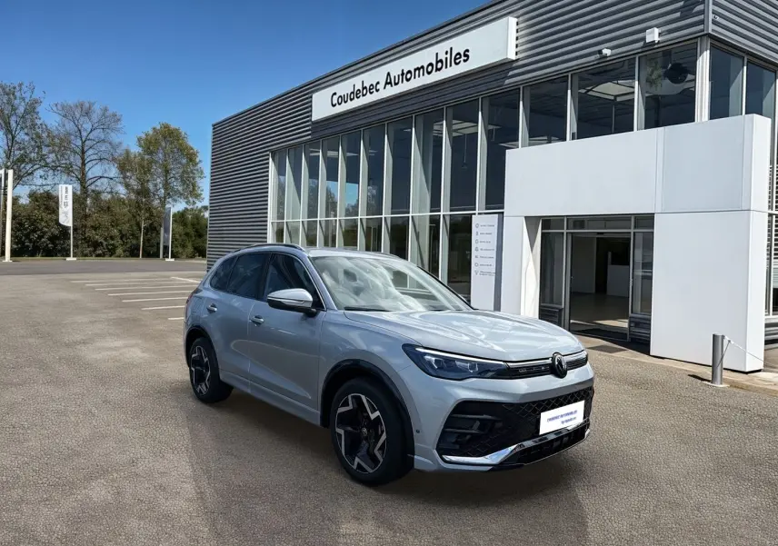 Volkswagen Tiguan gris en 3/4 avant droit, avec jantes noires brillantes et calandre moderne, devant un concessionnaire.