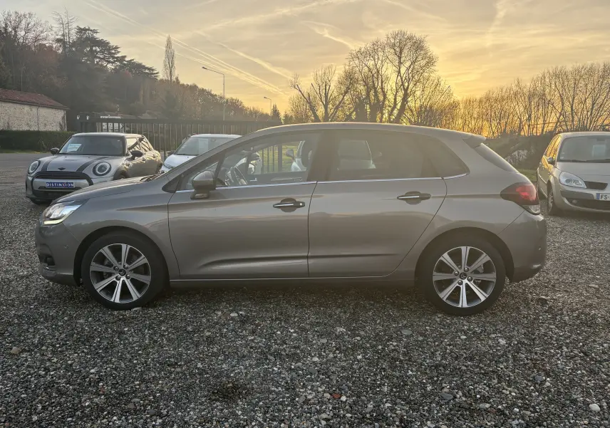 Profil côté gauche d'une Citroën C4 II gris métallisé de 2015, stationnée sur un sol gravillonné au coucher du soleil.
