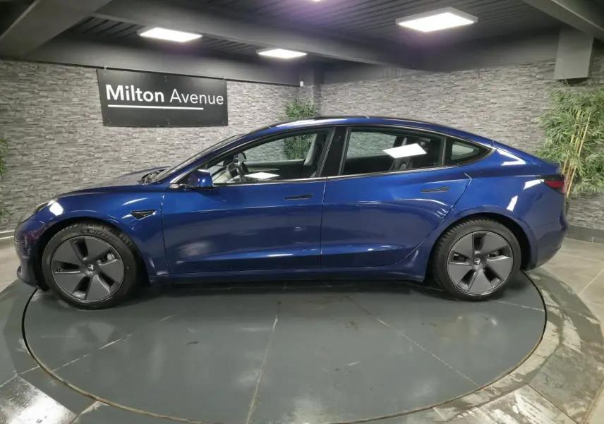 Profil côté gauche d'une Tesla Model 3 RWD 2021 bleu foncé avec jantes 18 pouces en aluminium dans un showroom.