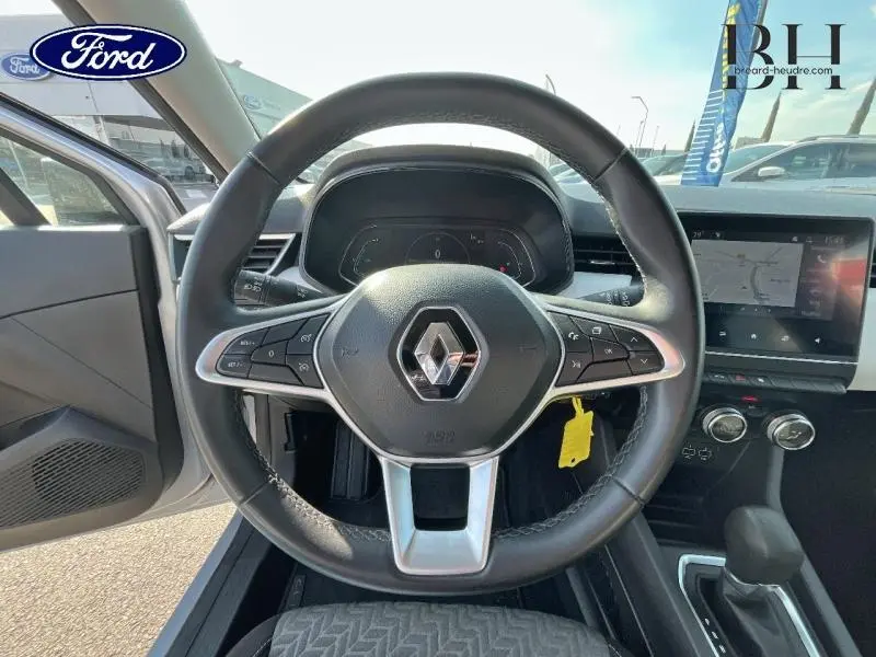Vue centrée sur le volant noir de la Renault Clio gris platine, avec tableau de bord digital et écran tactile GPS visible.