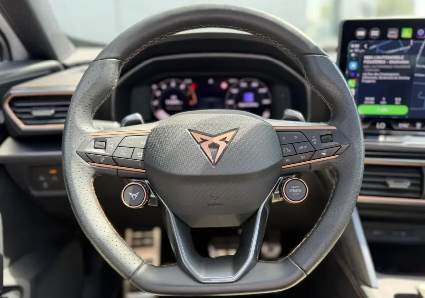 Volant Supersports CUPRA en cuir perforé avec logo cuivre, boutons satellites et tableau de bord numérique en arrière-plan.