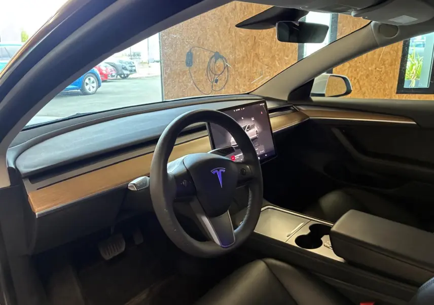 Vue intérieure côté conducteur du Tesla Model 3 blanc 2023, avec tableau de bord en bois clair et écran tactile central.