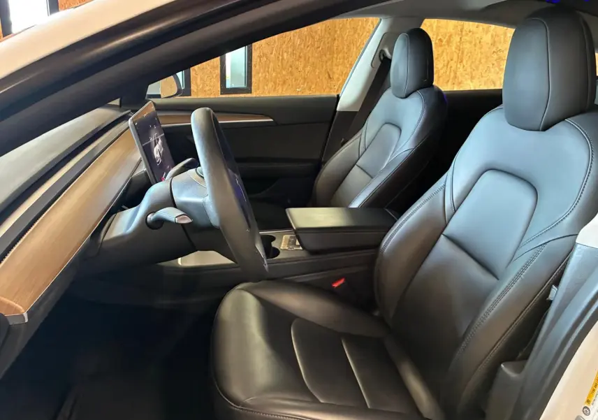 Vue intérieure côté conducteur de la Tesla Model 3 blanche 2023, sièges en cuir noir et tableau de bord avec écran tactile central.