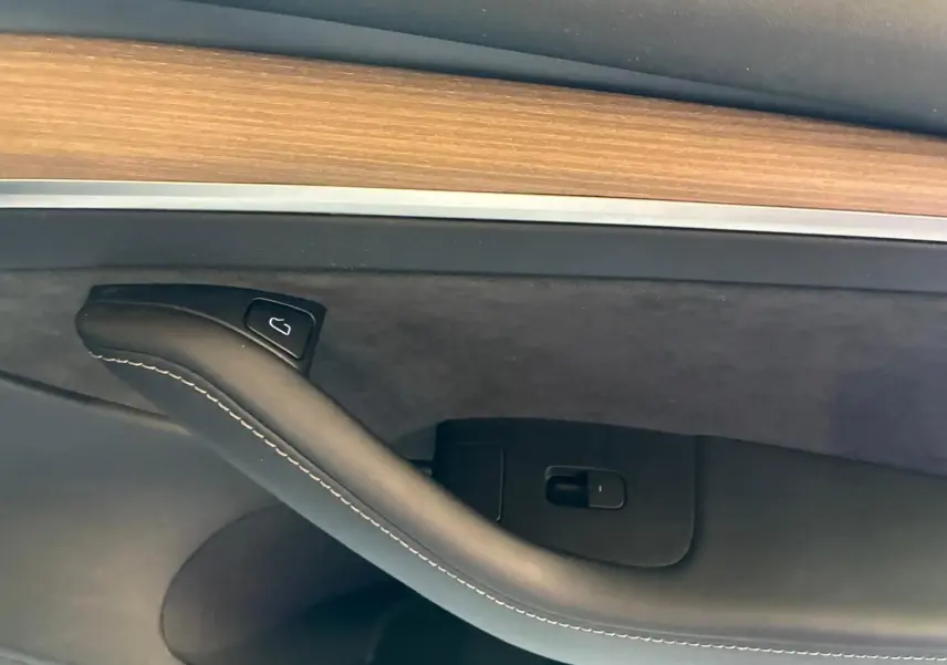 Détail de la poignée intérieure côté gauche d'une Tesla Model 3 blanche 2023 avec insert bois et surpiqûres blanches.
