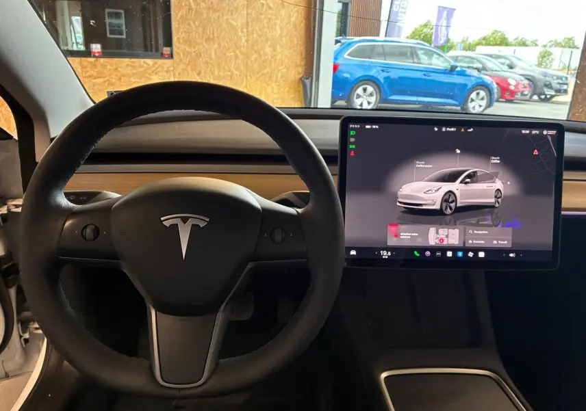 Vue intérieure centrée sur le volant noir et l'écran tactile du tableau de bord d'une Tesla Model 3 blanche de 2023.