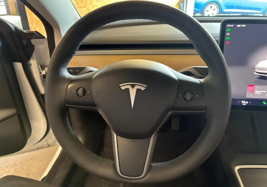 Vue intérieure centrée sur le volant noir d’un Tesla Model 3 blanc 2023, avec écran tactile visible à droite.