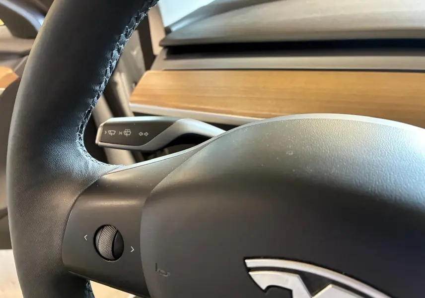Gros plan sur le volant noir de la Tesla Model 3 blanc, avec commande multifonction et tableau de bord en bois visible.