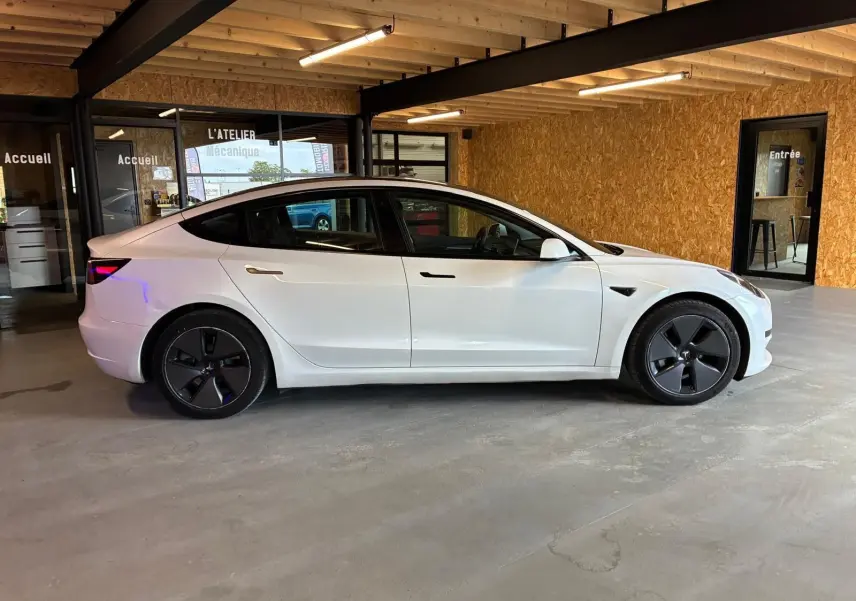 Vue de profil côté gauche d'une Tesla Model 3 blanche RWD 2023, avec jantes noires distinctives, dans un garage.