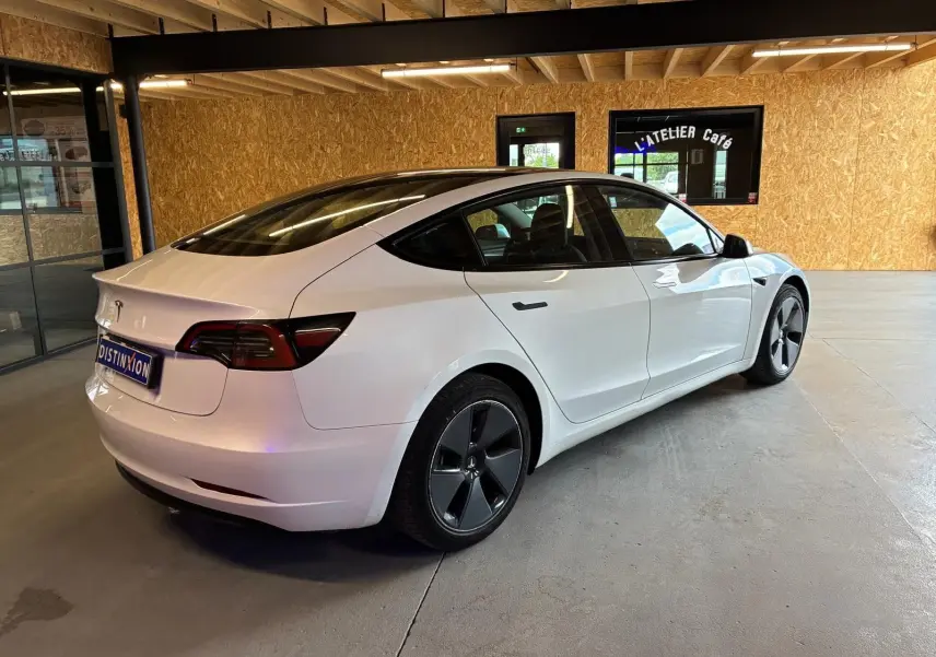 Tesla Model 3 blanc vue 3/4 arrière droit dans un garage avec jantes noires distinctives et toit panoramique.