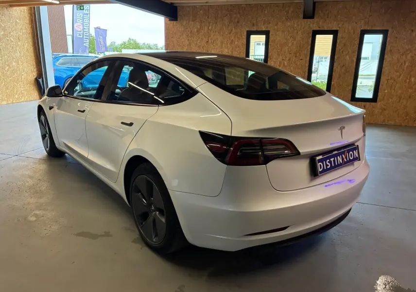 Tesla Model 3 blanc vue 3/4 arrière droit dans un garage, avec toit panoramique et jantes noires distinctives.