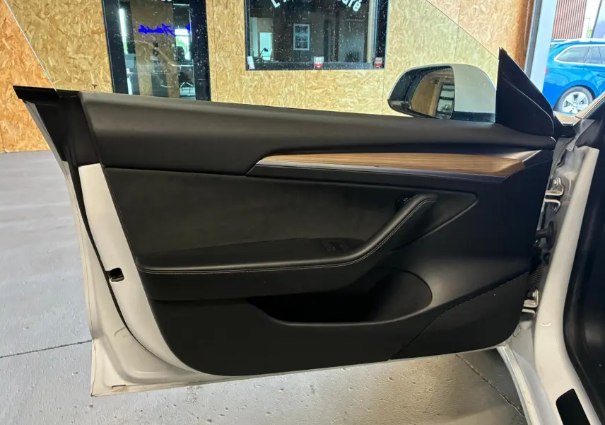 Vue intérieure de la porte côté gauche d’un Tesla Model 3 blanc 2023, avec garniture noire et insert imitation bois.