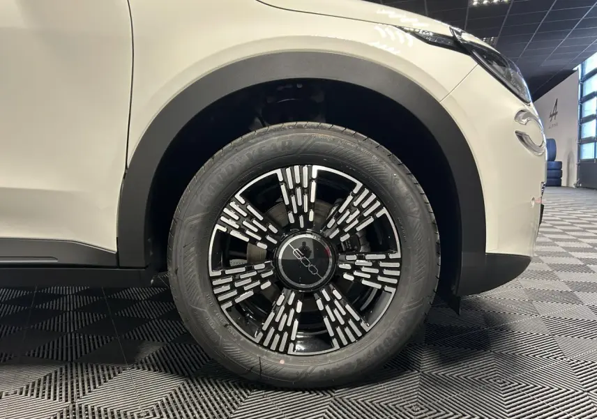 Gros plan sur la roue avant droite noire et chrome d'une FIAT 600 blanche, avec partie avant du véhicule visible.