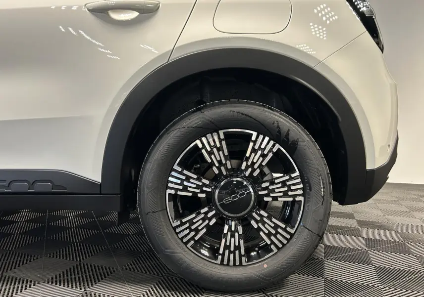 Gros plan sur la roue arrière droite noire et argentée du Fiat 600 blanc, avec passage de roue et partie de la carrosserie visibles.